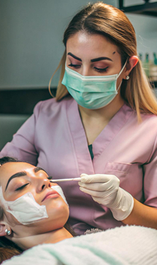 Esteticista con mascarilla quirúrgica aplicando tratamiento facial con pincel a una paciente relajada, cuidado y seguimiento postoperatorio en clínica de rinoplastia Alicante