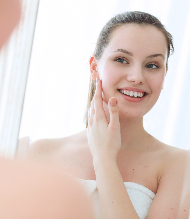 Mujer joven sonriente mirándose al espejo y tocándose el rostro, transmitiendo satisfacción con su nariz y rasgos faciales tras una rinoplastia